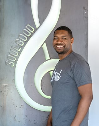 Andrew Osakue, partial owner and operator of Soul Good, stands in front of neon-lit sign