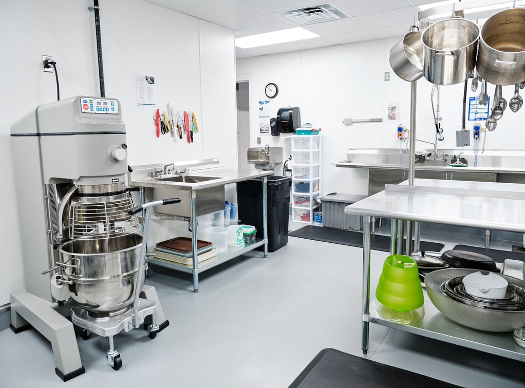 A brightly-lit kitchen contains a commercial floor mixer, sink, and work table