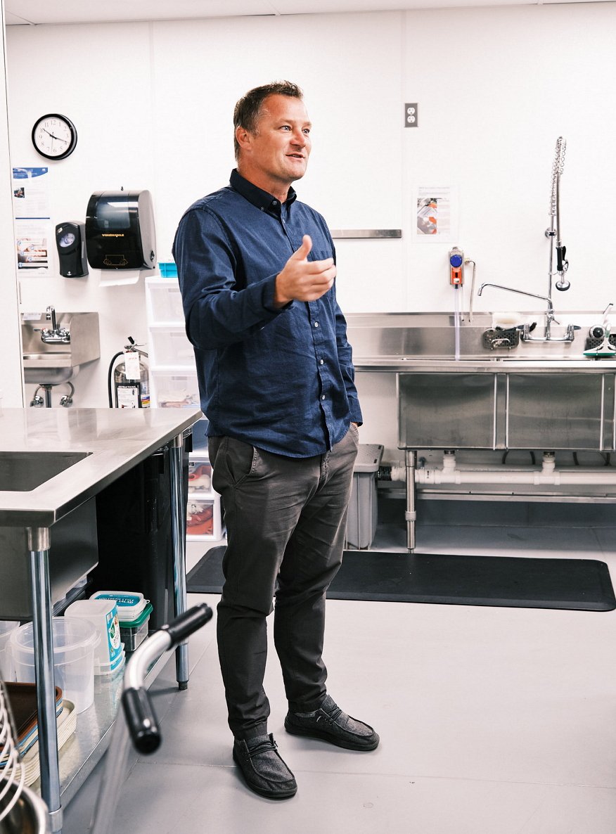 Pastor Cain is standing in a kitchen wearing a blue shirt and grey pants. Equipment is visible in the foreground and background.