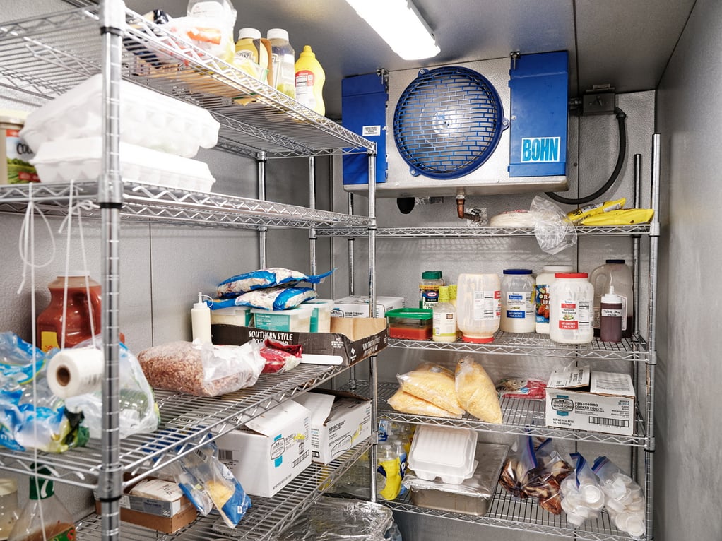 Packages and jars of food are arranged on wire shelving inside a walk-in cooler
